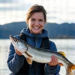 Porträt von Claudia Müller, einer freundlichen Angelführerin, die einen gefangenen Hecht stolz in die Kamera hält, vor dem Hintergrund des Zugersees.
